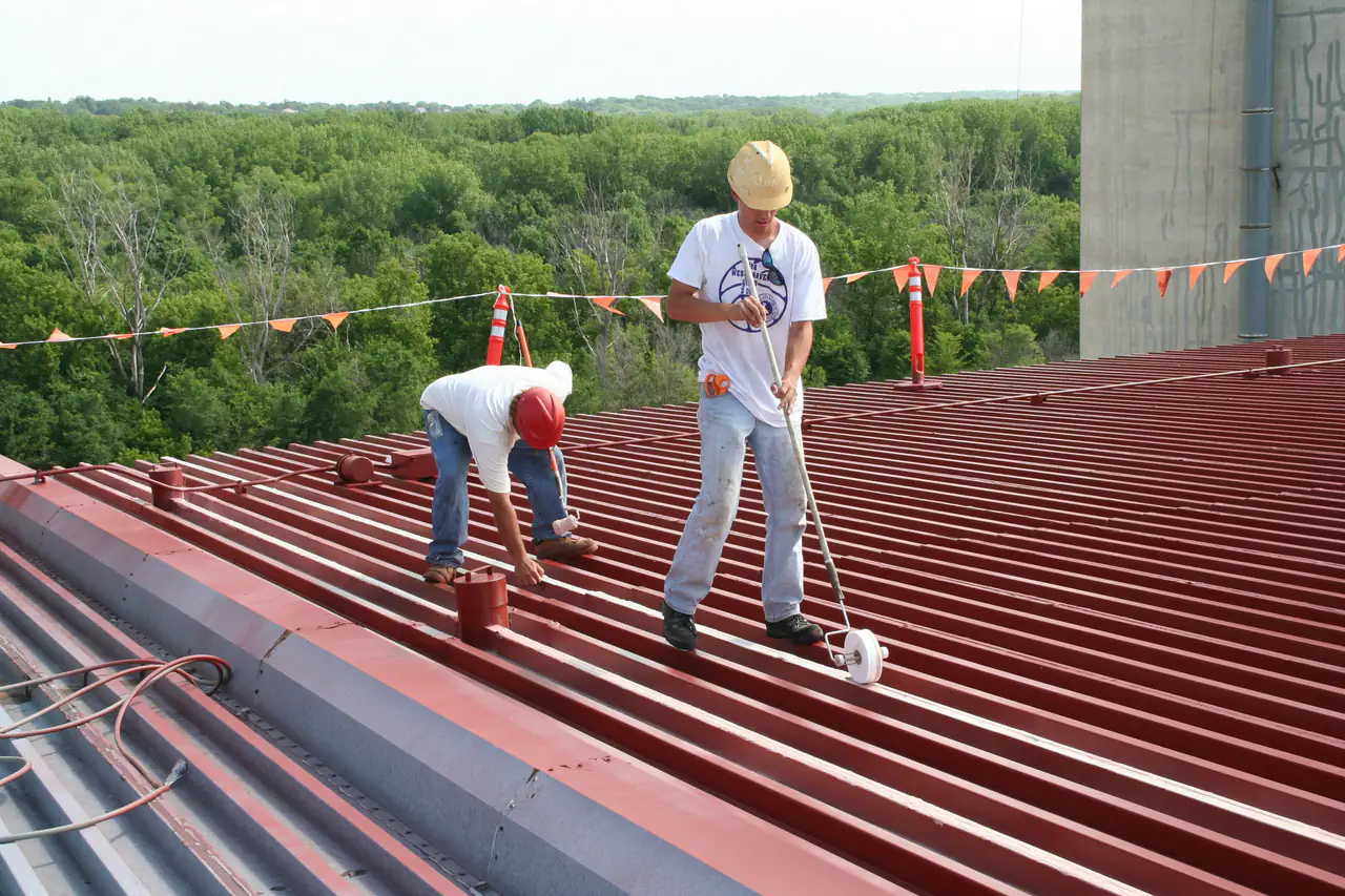 Two roofers rolling coating onto a red standing-seam metal roof