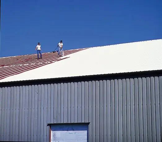 A red metal building roof partly covered in white coating, with two roofers working