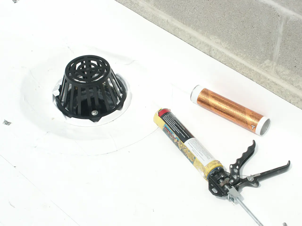 A roof drain set into a white membrane roof, with a tube of sealant and a caulk gun beside it