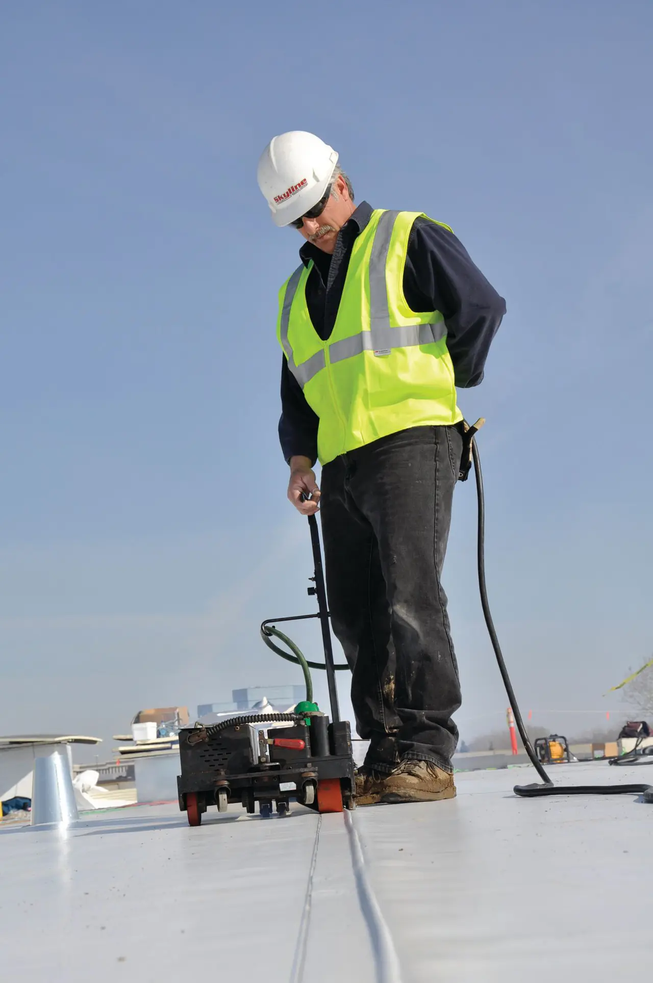 A roofer in a hard hat and safety vest heat-welding the seam of a white single-ply roof