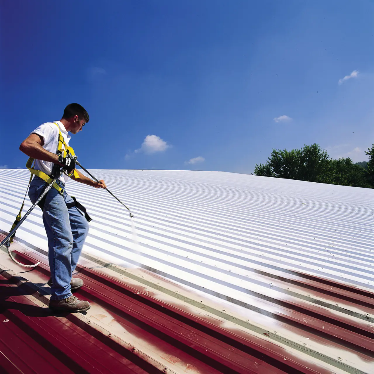 A roofer in a safety harness spraying white coating over a faded red metal roof